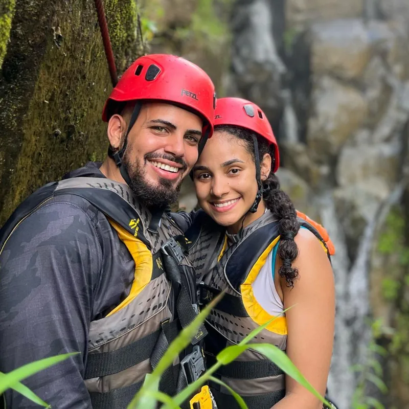 Couple in red climbing helmets and harnesses with a waterfall behind them, via ferrata, El Yunque, Puerto Rico